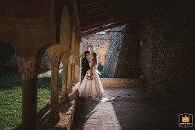 A newly married couple is captured in a sun-drenched, romantic portrait, standing amidst the historic architecture and warm light of the ancient Tuscan location.